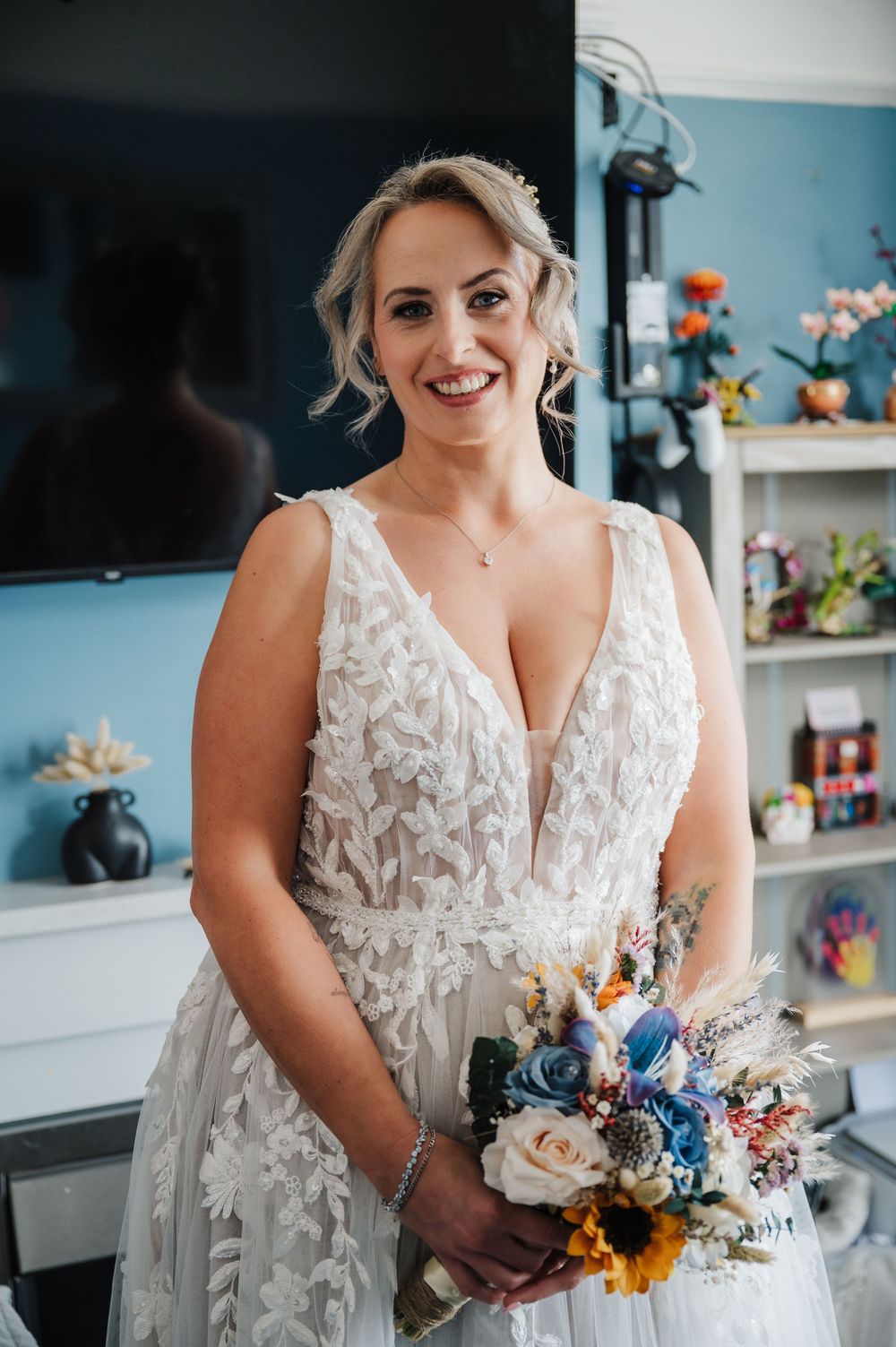 Smiling bride in a lace V-neck wedding gown holding a colorful bouquet with sunflowers and blue roses.