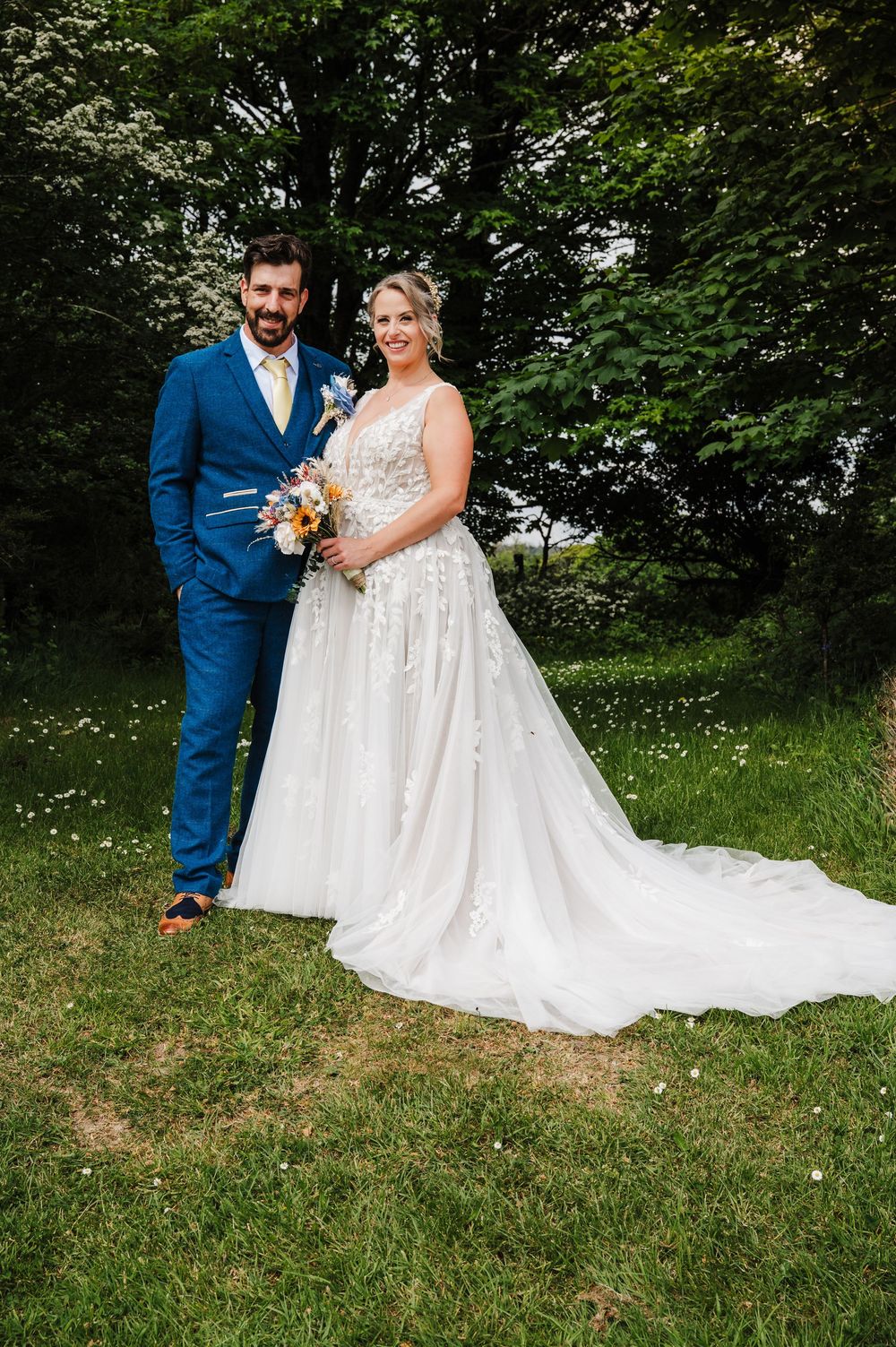Bride and groom pose together outdoors in lush garden, bride in floral wedding gown.