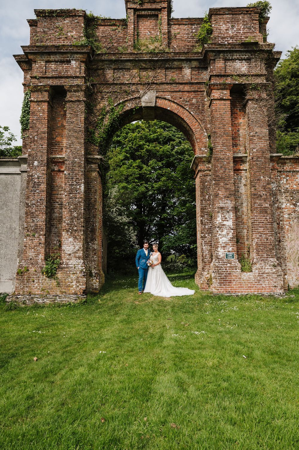Bride and groom posing under a grand brick archway surrounded by lush greenery at a wedding venue.