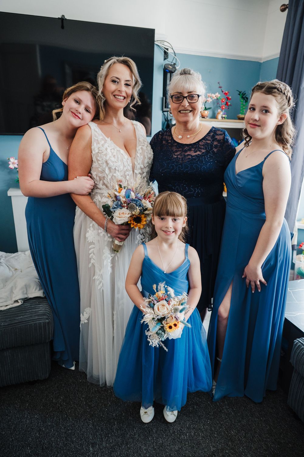 Bride in white gown with bridesmaids and flower girl in blue dresses holding bouquets with sunflowers.