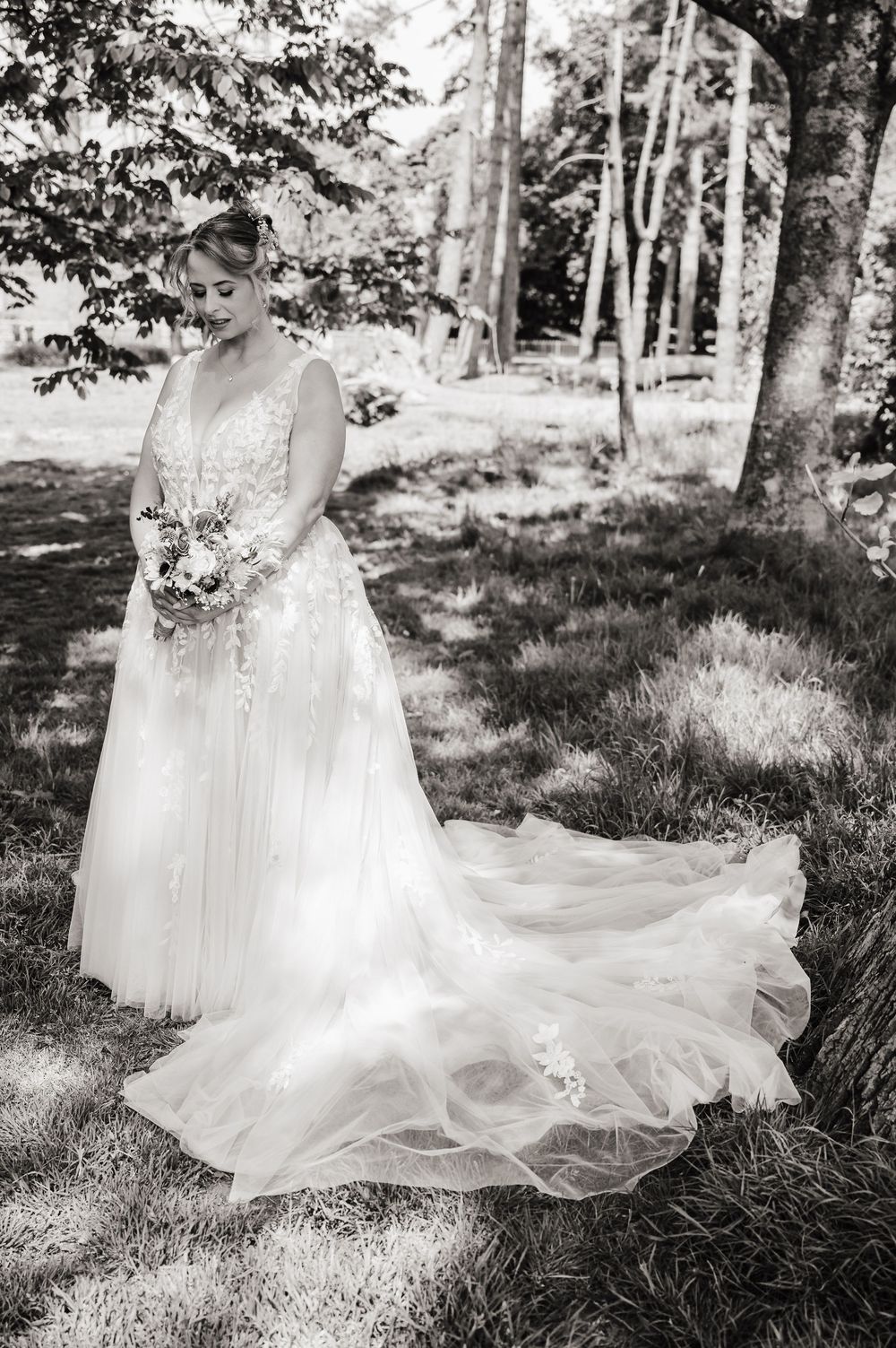 Bride in elegant lace wedding gown with flowing train holds bouquet outdoors in black and white portrait.