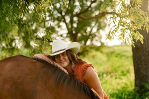 A rider in a cowboy hat and orange top leans over their horse's back in golden sunlight.