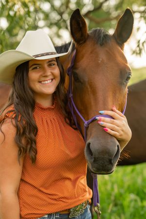 A western-styled rider in orange stands affectionately with their chestnut horse wearing a purple halter.
