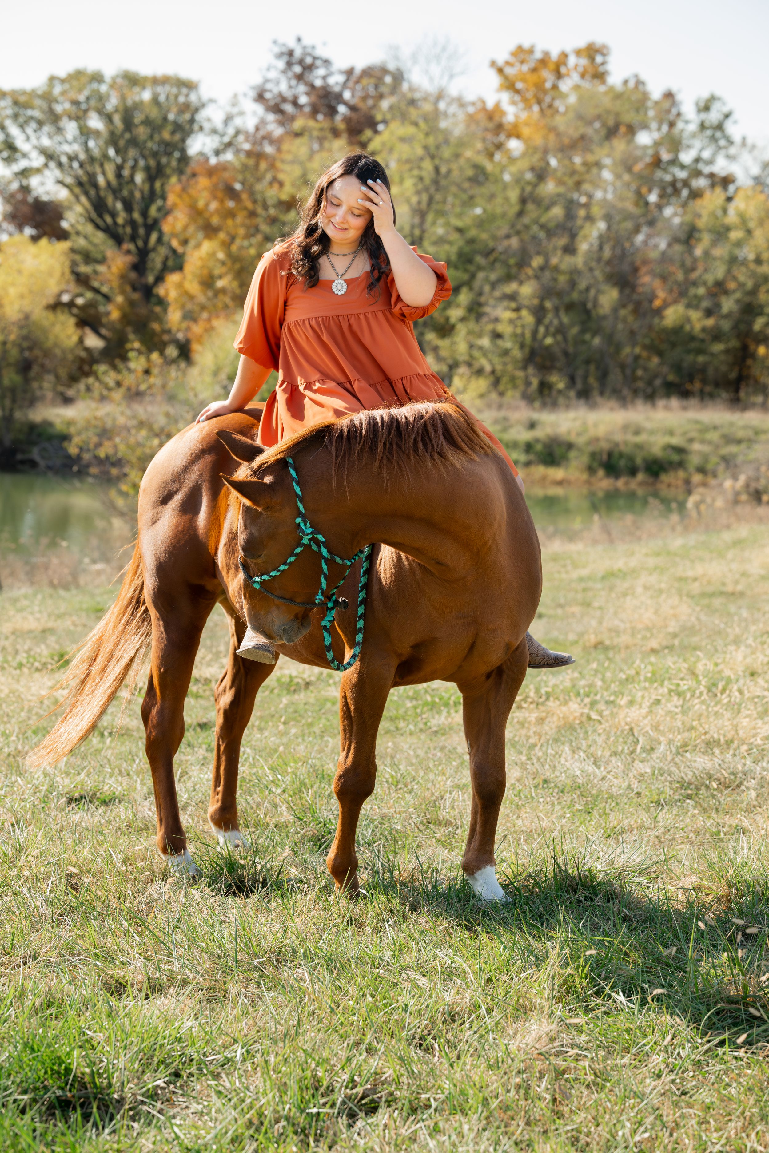 Rider in orange top aboard chestnut horse with turquoise reins in fall meadow.