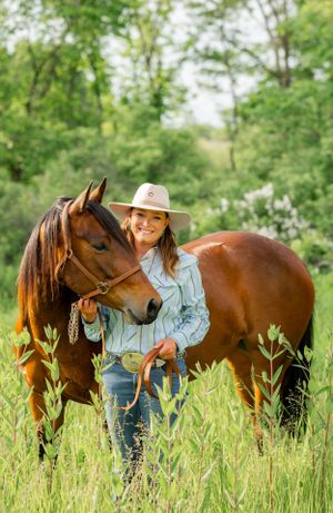A beautiful bay horse stands with a rider in a light denim shirt and cowboy hat in a lush green field.