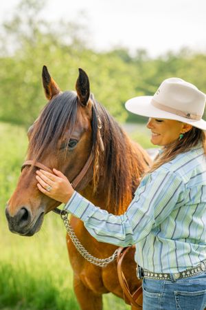 A rider in western attire gently pets a bay horse's face in an outdoor summer setting.