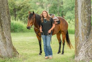 A bay horse stands with a rider in casual western wear between two trees in a green field.