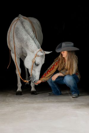 A white horse lowers its head while being photographed with someone in western attire.