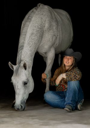 A dappled white horse bends down to graze next to someone in western wear and cowboy hat.