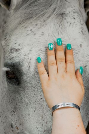 A hand with turquoise nail polish touches a white horse's dappled face while wearing a silver bracelet.