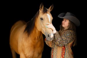 A palomino horse standing against a black background being petted by someone wearing a cowboy hat and patterned top.