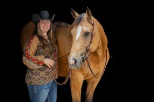 A chestnut horse poses with someone in western attire against a black background.