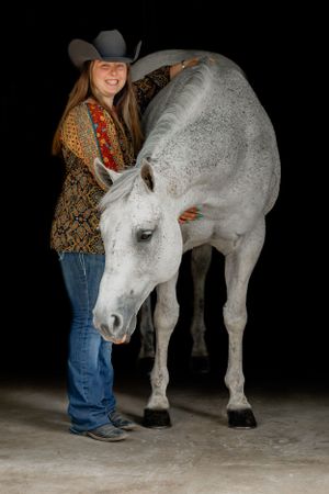A white dappled horse poses with a western-styled person against a black background.