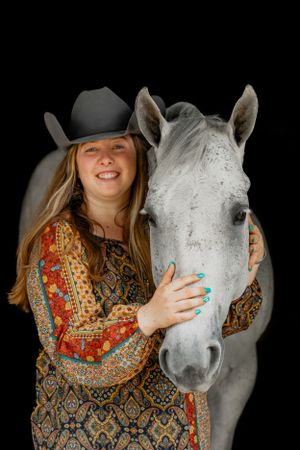 A white horse stands with someone wearing a patterned top and black cowboy hat against dark backdrop.