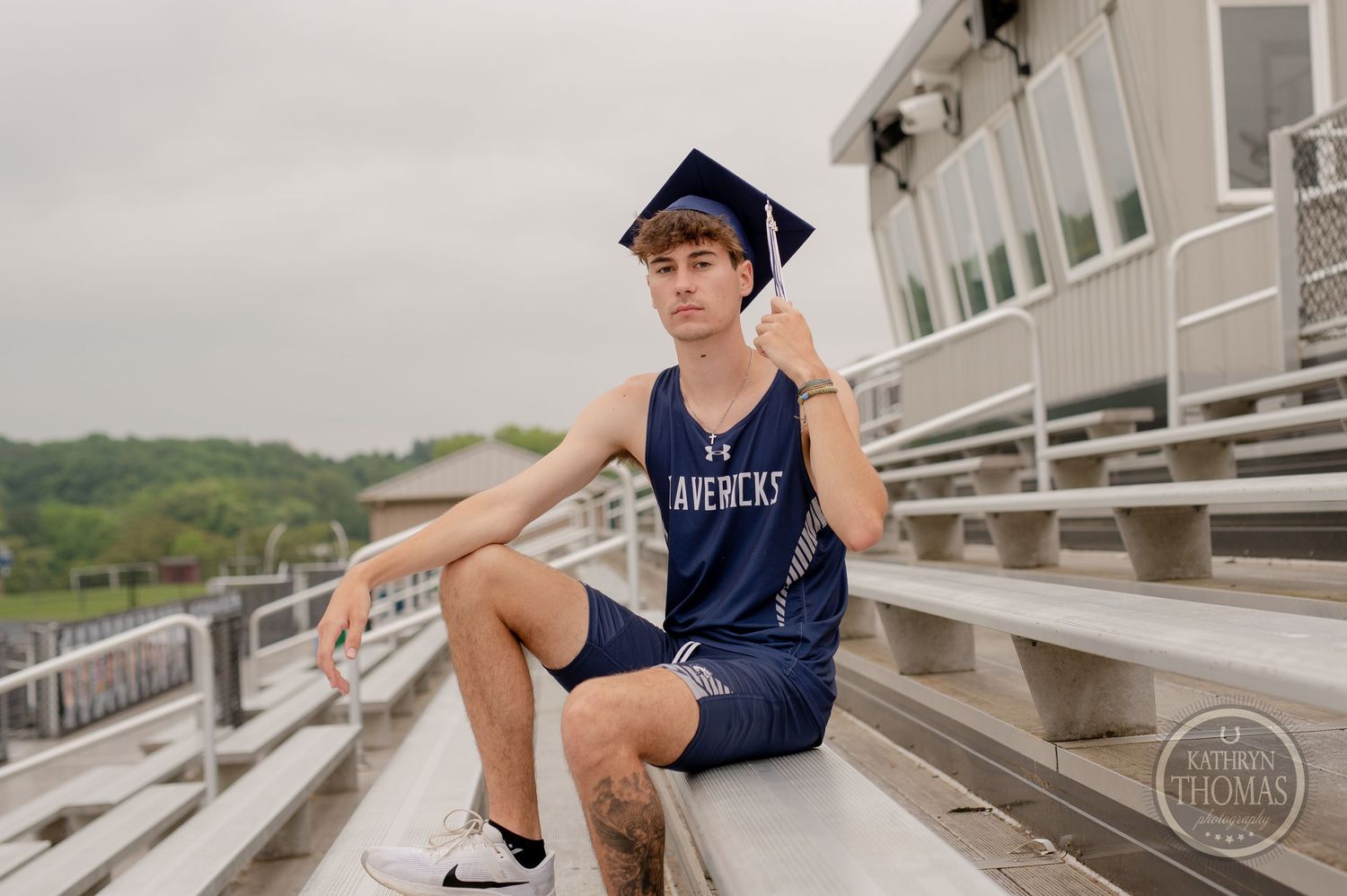 A person in graduation cap and navy uniform sits on aluminum bleachers at an athletic facility.