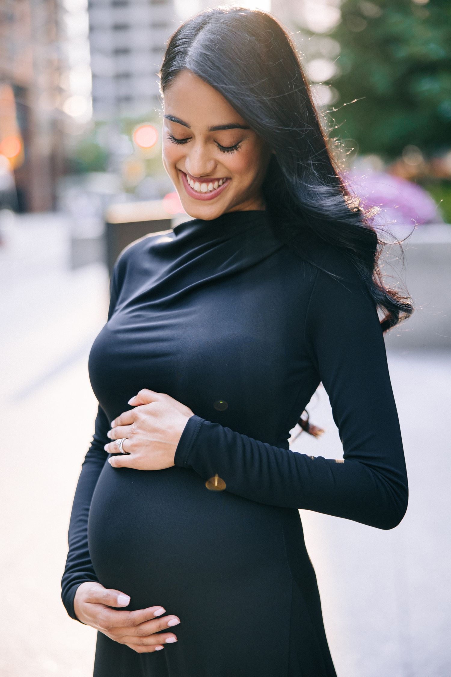 Person wearing a fitted black long sleeve dress caresses their pregnant belly while smiling with eyes closed.