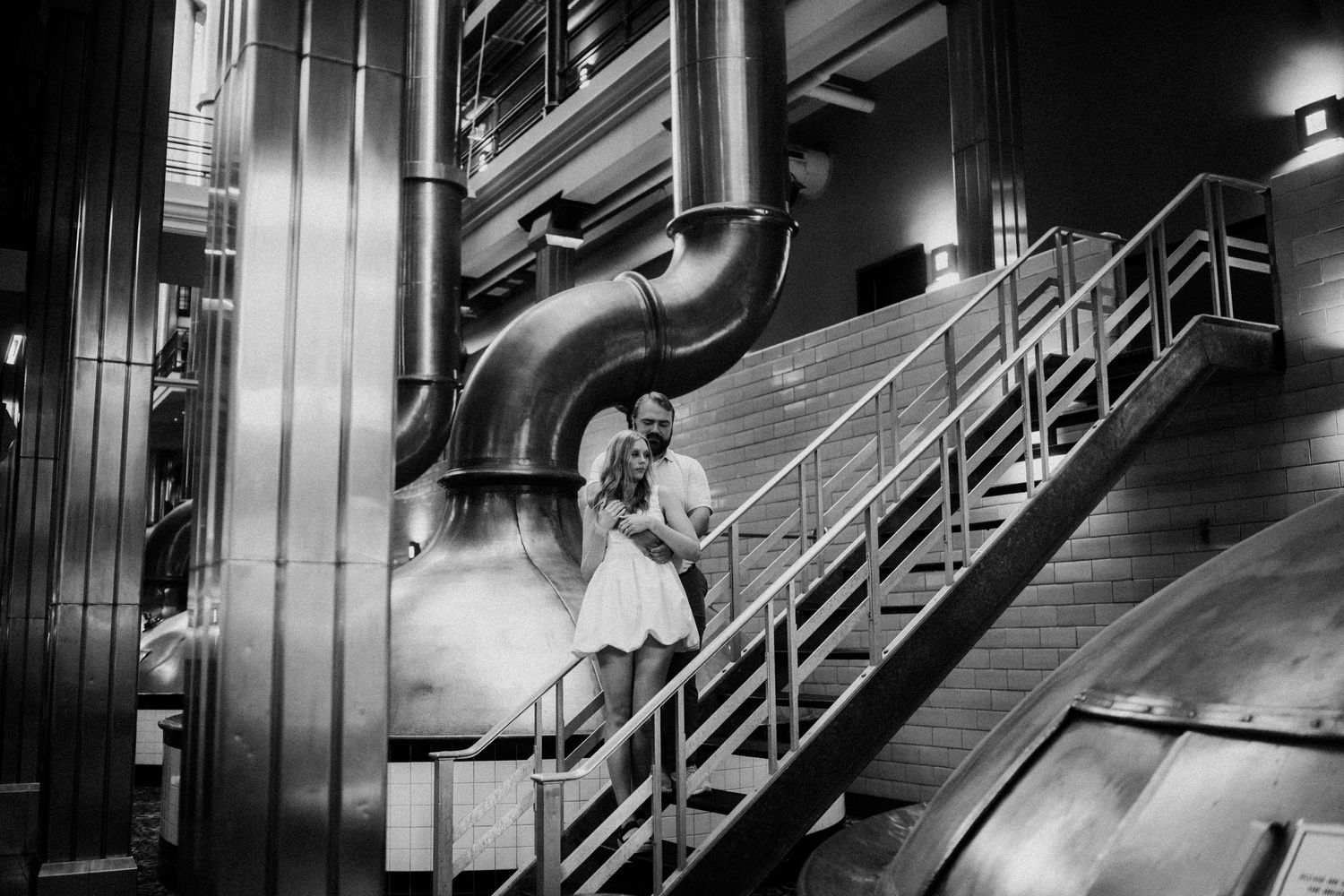 A black and white industrial interior with metal staircase and large pipes in a dramatic factory setting.