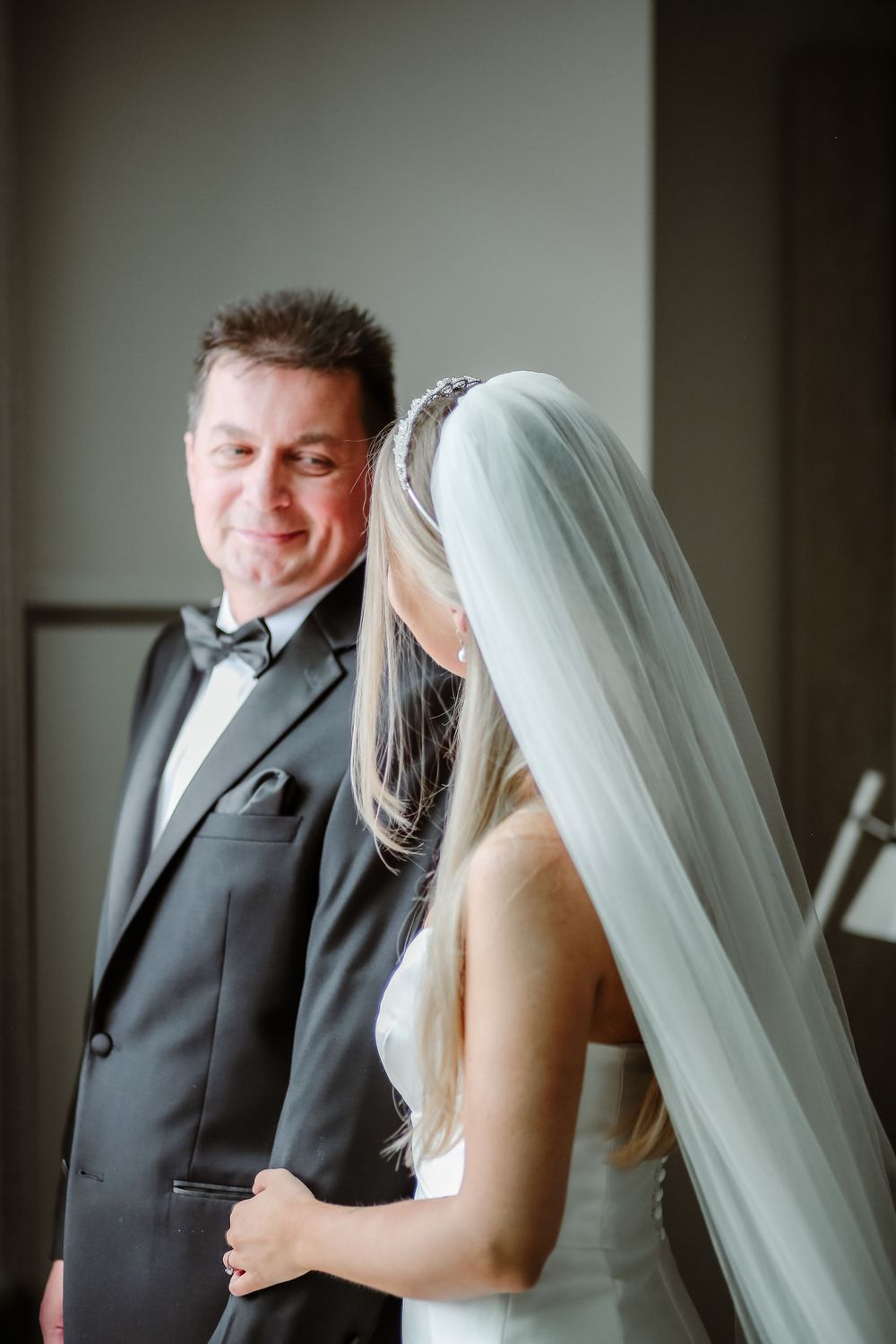 A bride in a white wedding dress and veil stands next to her companion in a black tuxedo during an intimate moment.