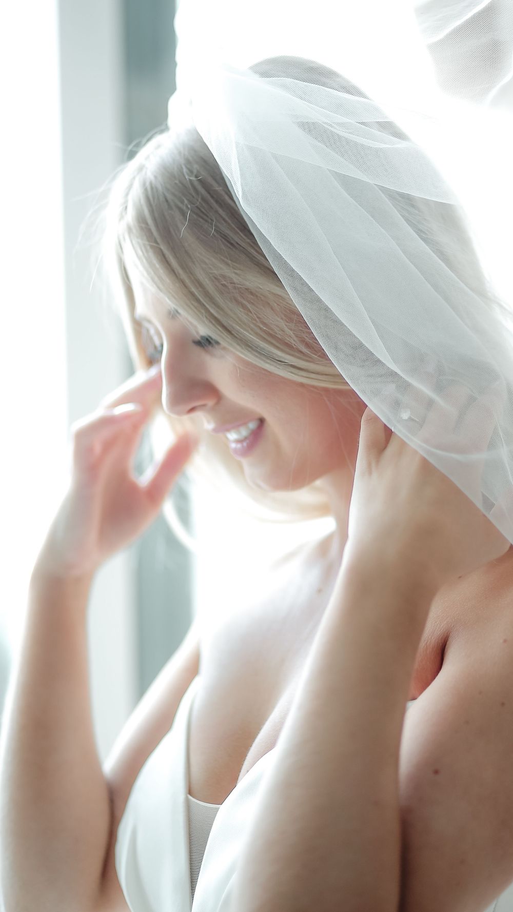 Close-up portrait of a glowing bride adjusting her delicate veil near a window.