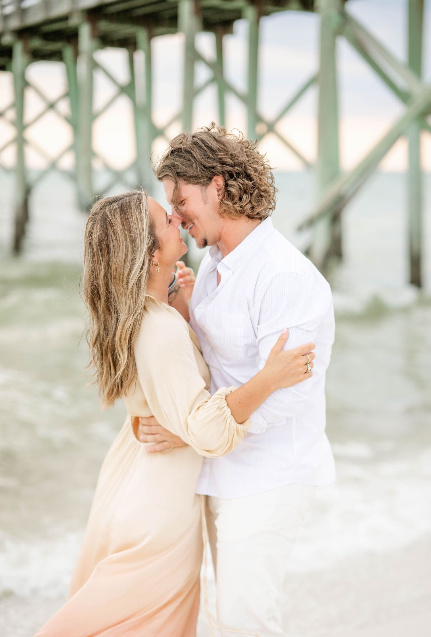 A romantic coastal engagement photo with a couple embracing near a pier in pastel peach and white outfits.