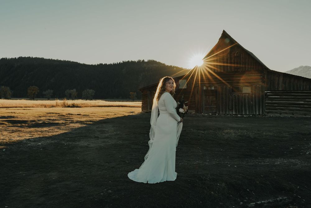Bride in white gown poses at sunset near rustic barn with mountain backdrop in Grand Teton National Park.