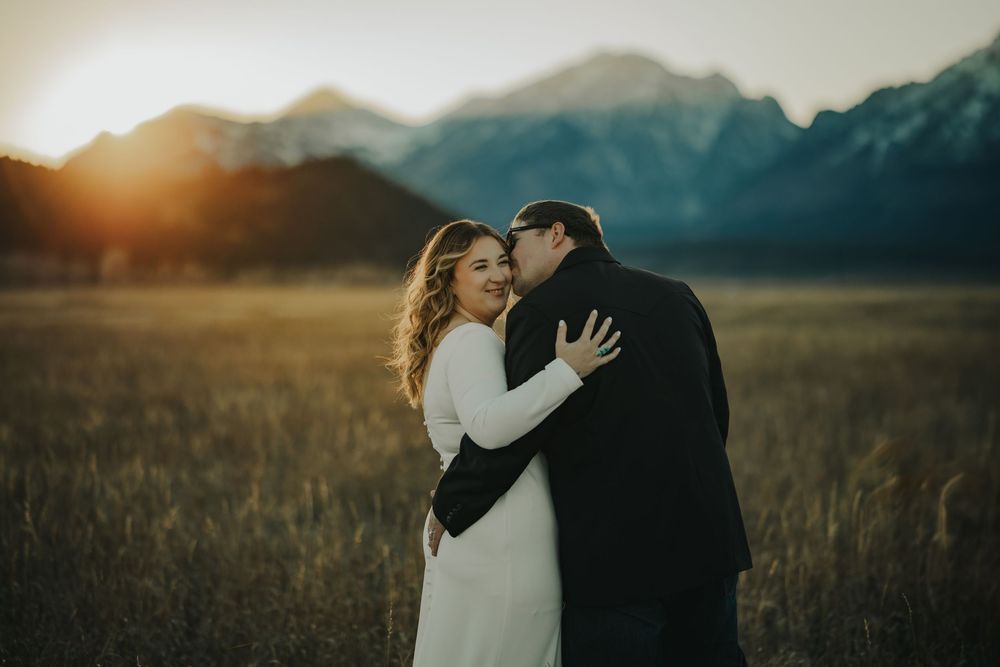 Couple embracing at golden hour in a mountain meadow, bride in white gown, groom in dark suit, sunset peaks behind them.
