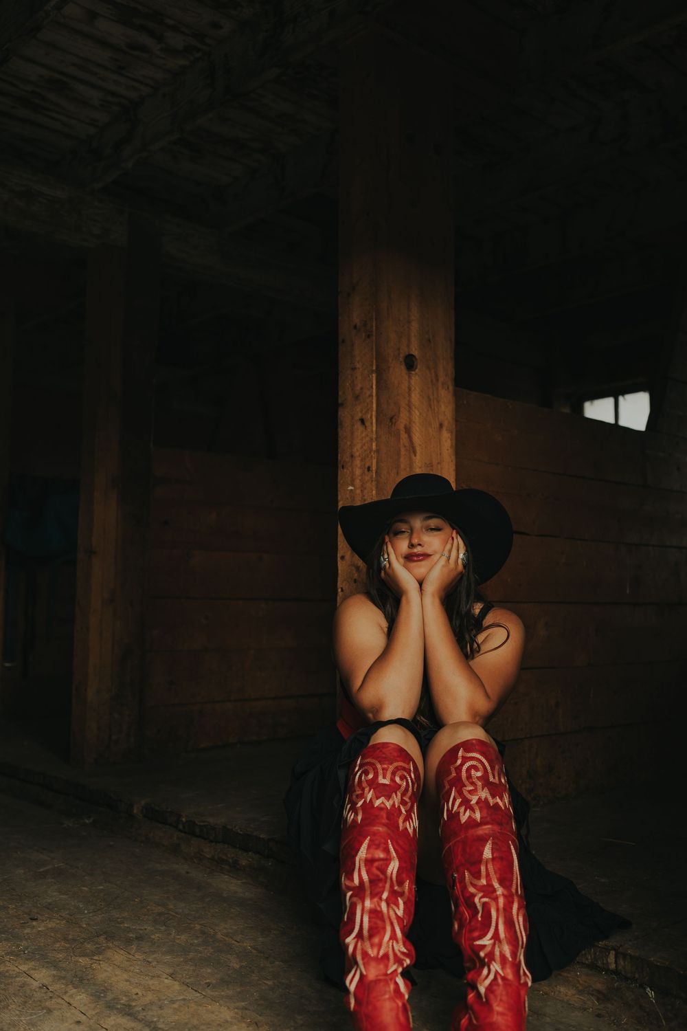 Woman in black cowboy hat and red boots sits against wooden barn post, smiling with hands clasped under chin.