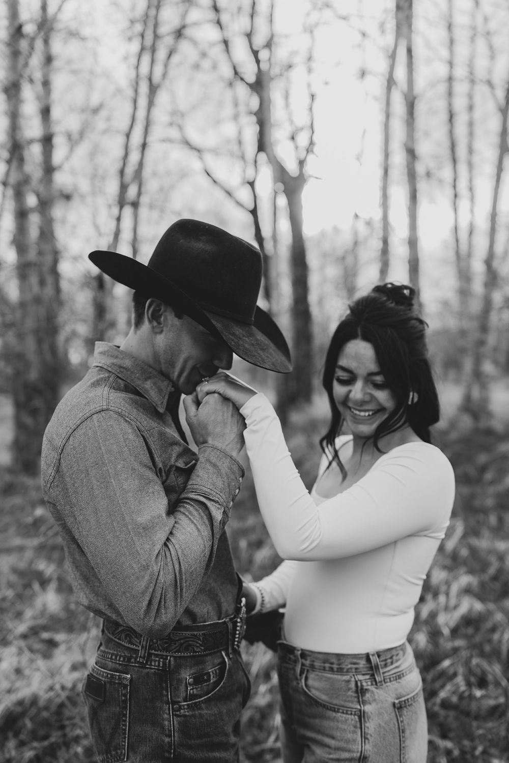 Black and white photo of a couple sharing a romantic moment outdoors, man in cowboy hat kissing woman's hand.