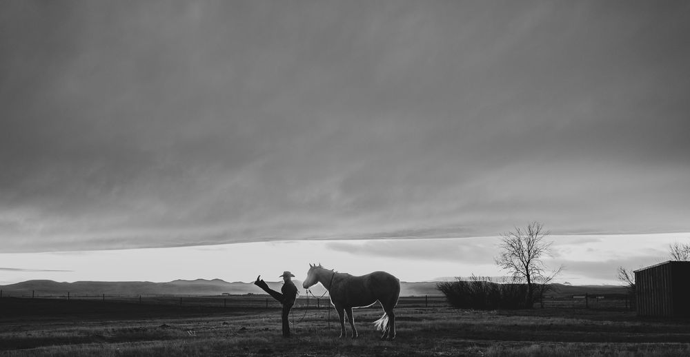 Black and white photo of a person standing beside a horse on an open ranch plain under dramatic cloudy skies.