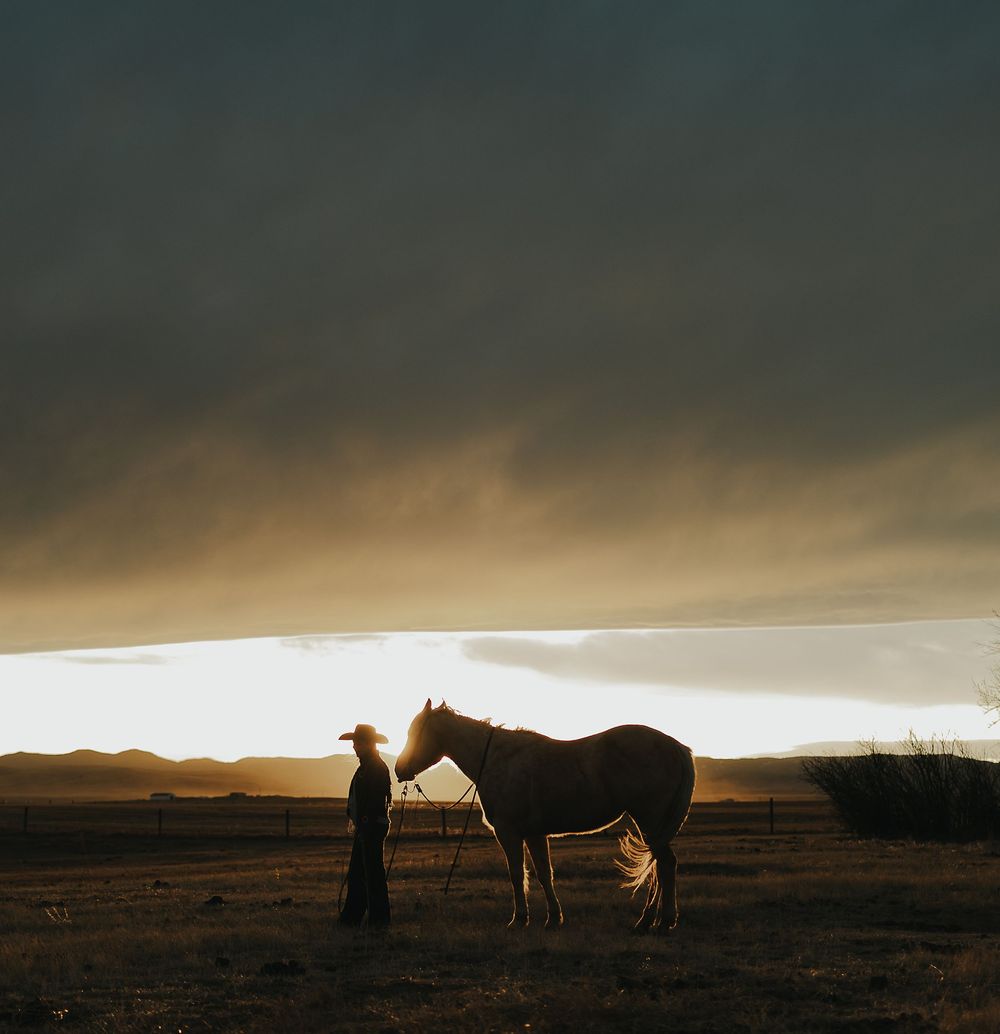 Cowboy standing with a horse at sunset on open plains under dramatic storm clouds.