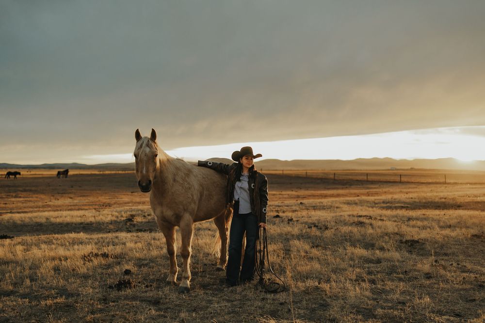 A cowboy in a hat stands beside a pale horse at golden sunset on a vast open prairie landscape.