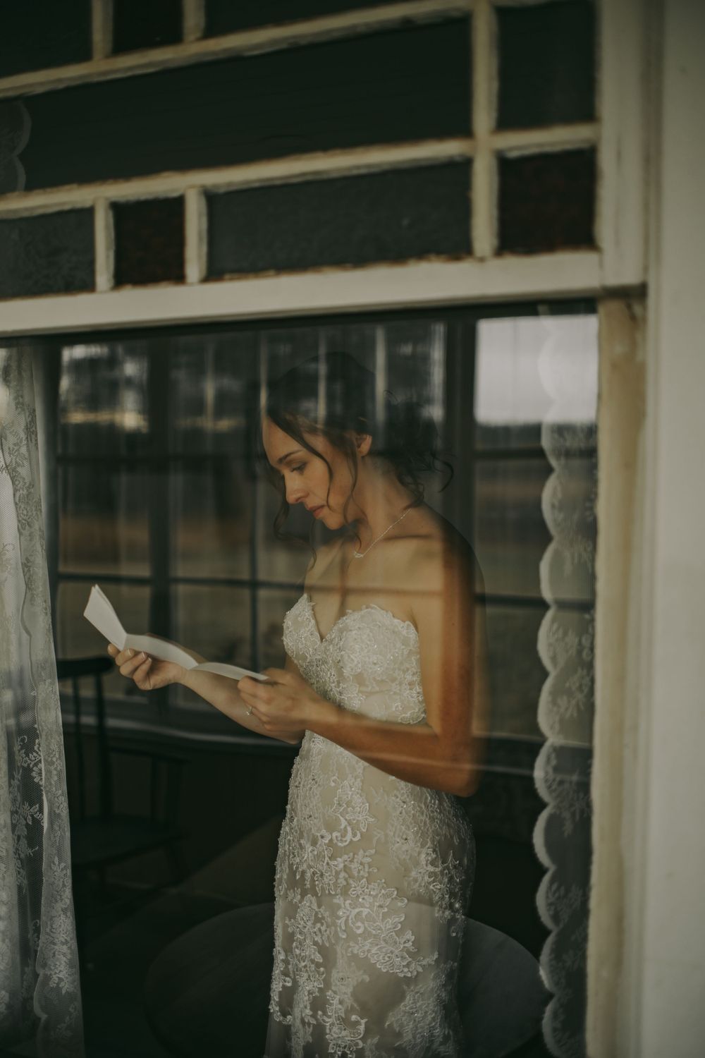 A person in a lace wedding dress reads a letter by a window.