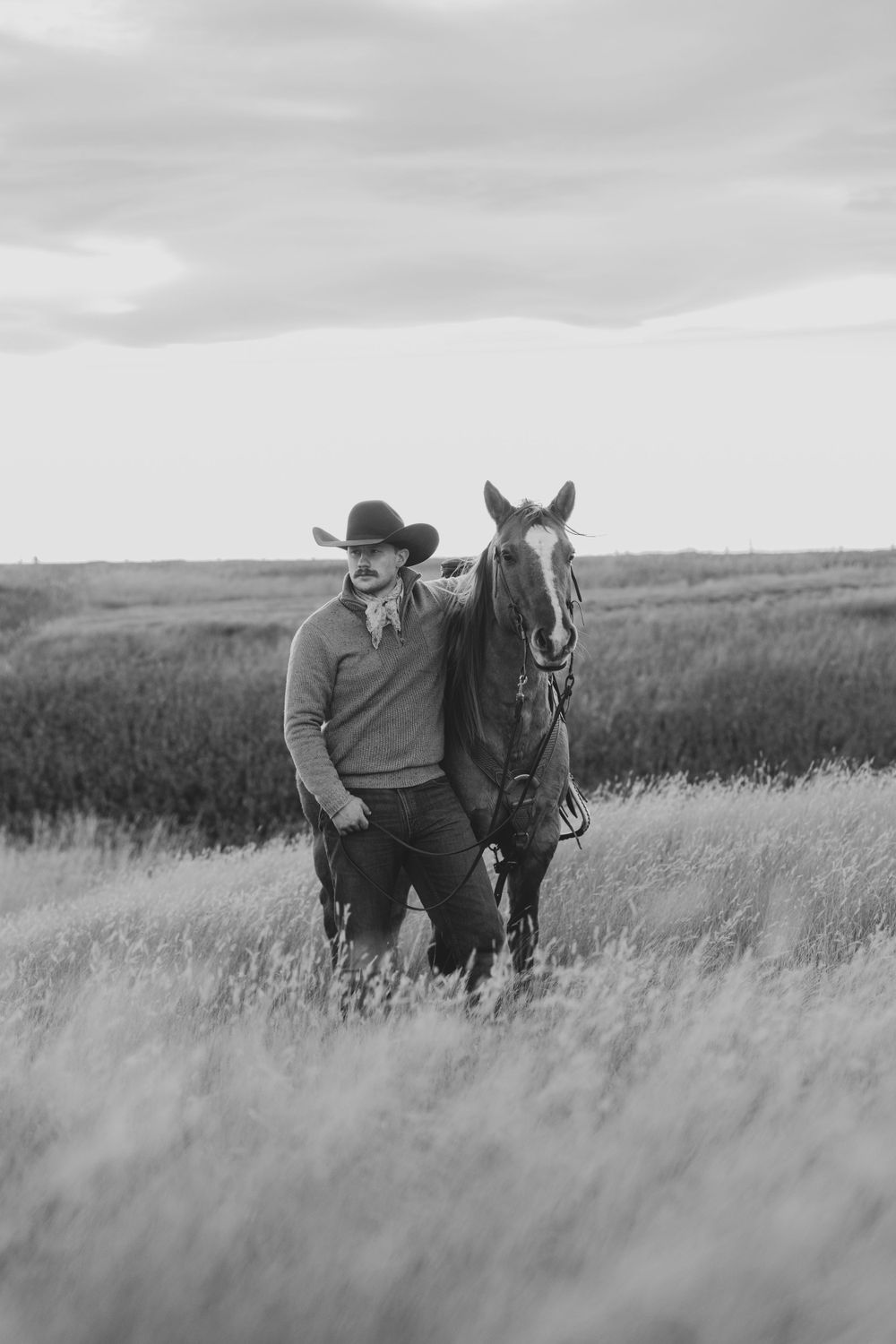 A person in western attire stands with their horse in tall prairie grass under cloudy skies.