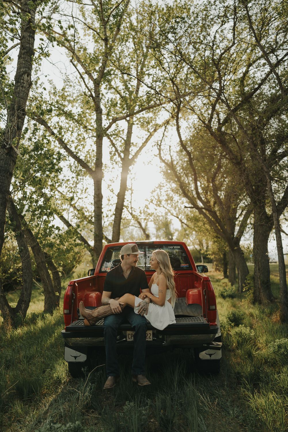 Couple sitting together on the tailgate of a red pickup truck surrounded by sunlit trees in a rustic outdoor setting.