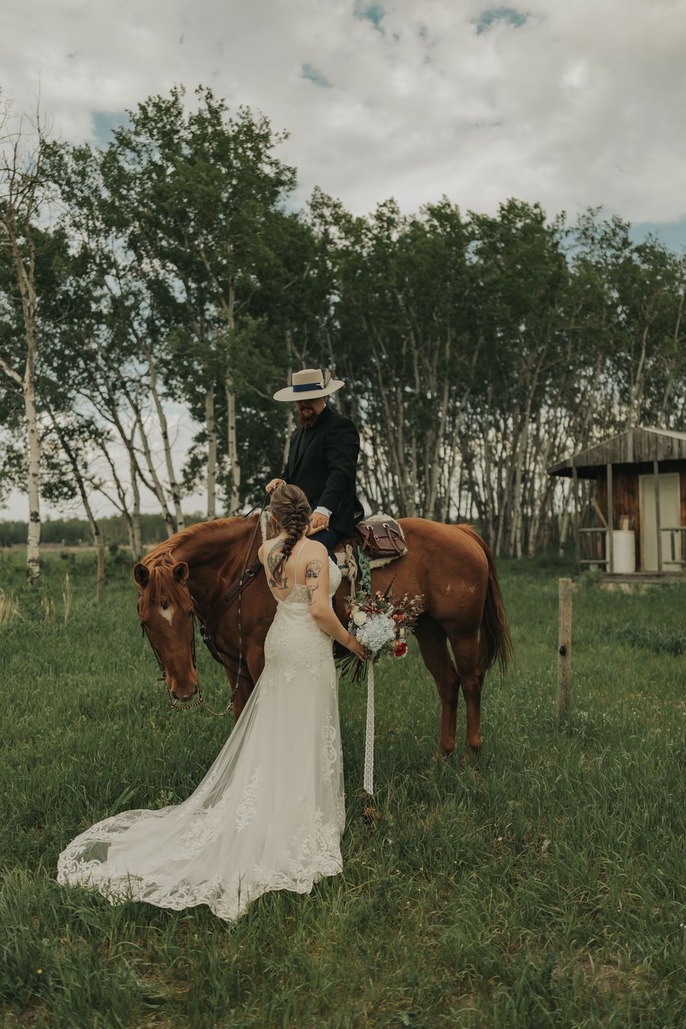 Bride in lace gown holds bouquet beside groom on chestnut horse at rustic outdoor wedding ceremony.