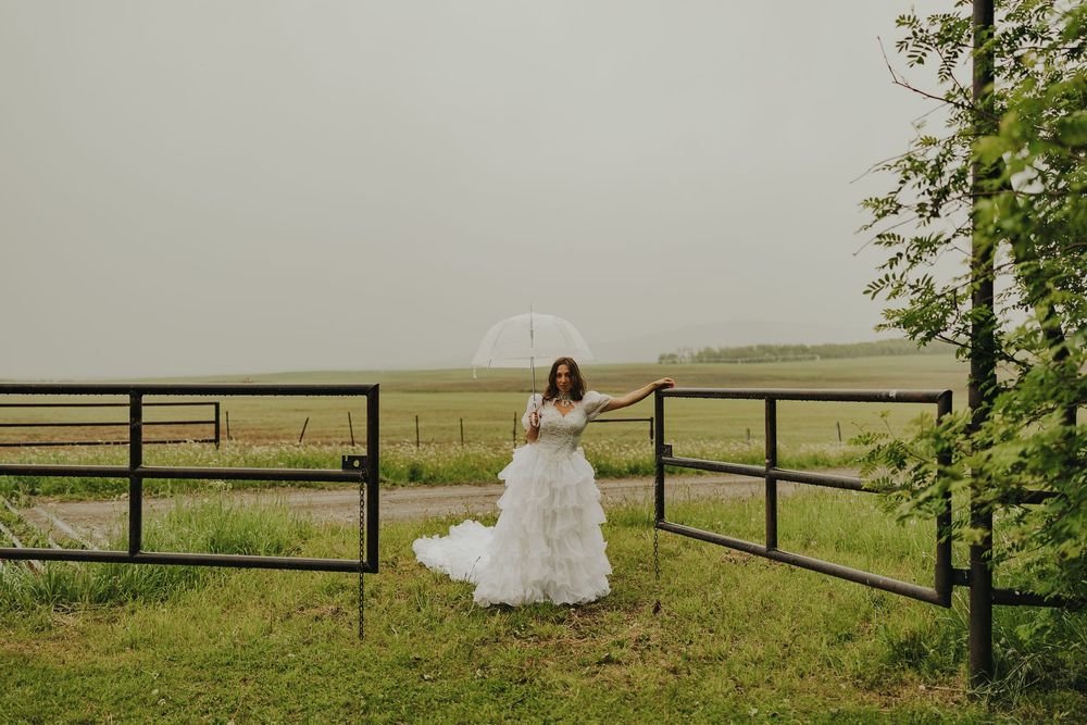 Bride in white gown holding clear umbrella by farm gate on a rainy day in a green countryside.