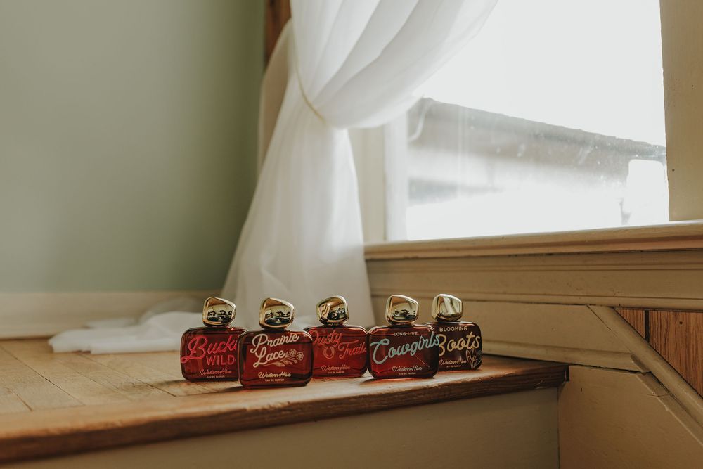 Four red and gold perfume bottles arranged on a windowsill with sheer white curtains in soft natural light.