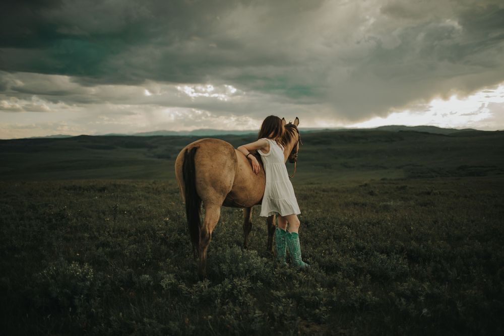 A person in a white dress and turquoise boots stands beside a horse in a stormy field.