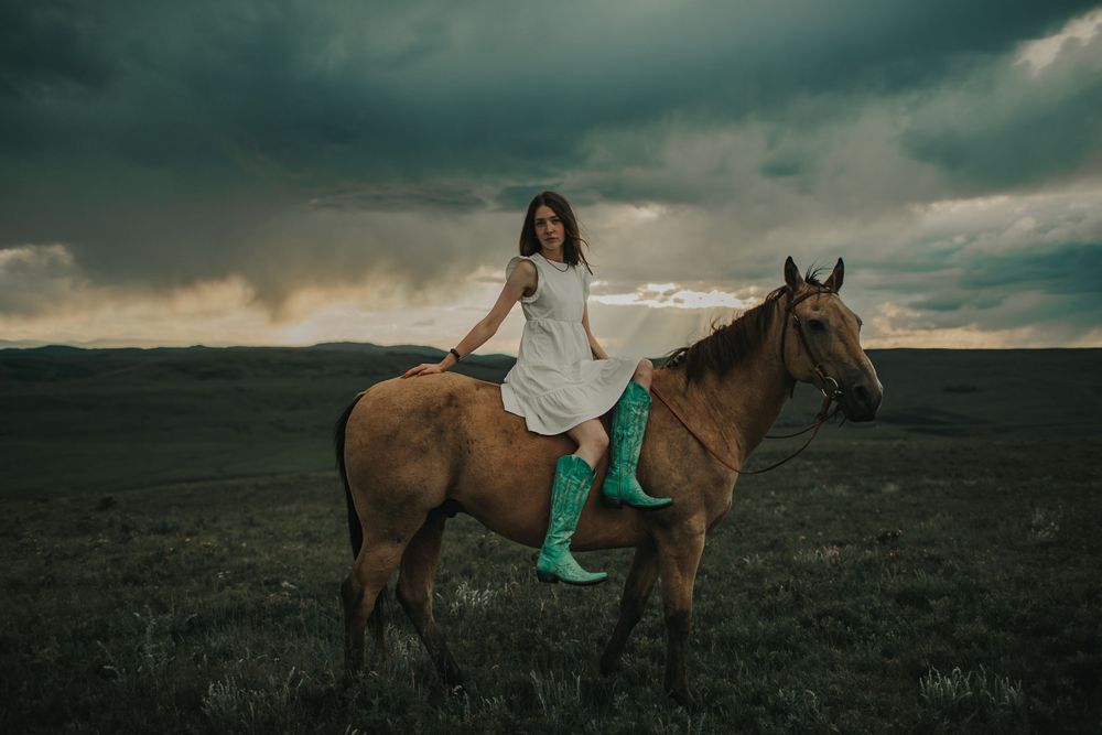 A person in turquoise boots rides horseback across a dramatic stormy landscape.
