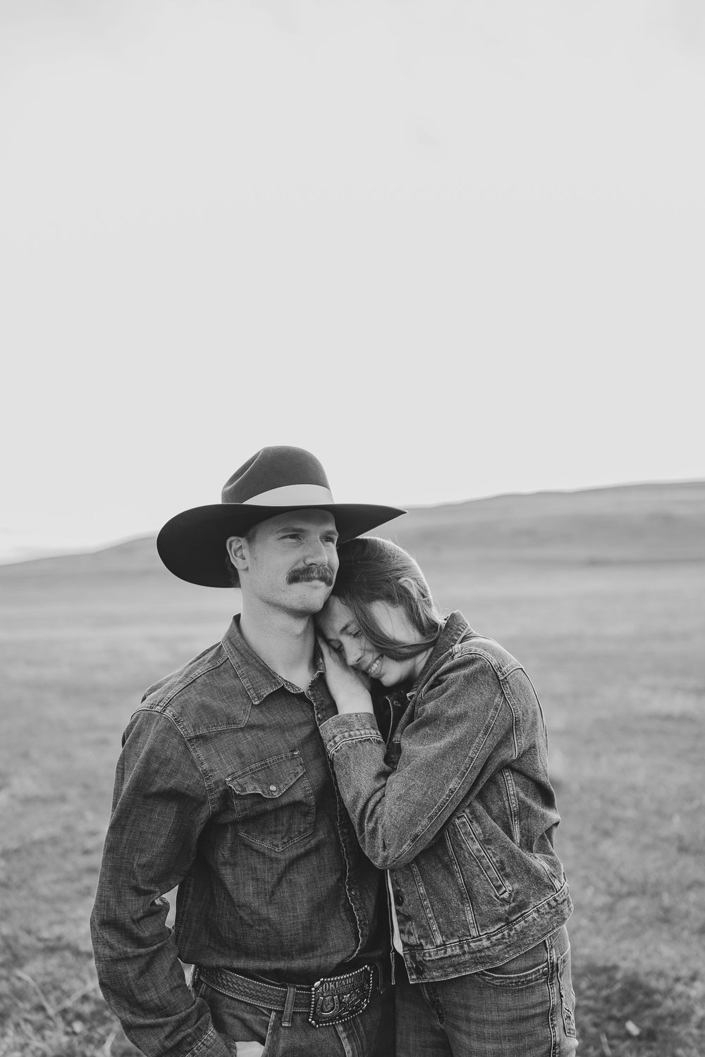 Black and white photo of a couple in denim jackets, man in cowboy hat, woman resting head on his shoulder outdoors.