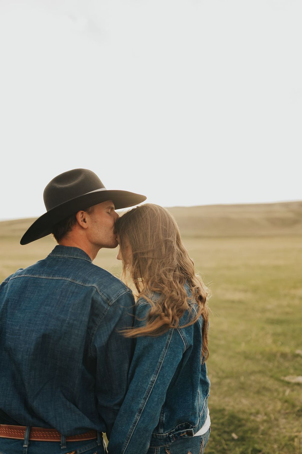 A couple in denim jackets shares a tender kiss in an open field, man wearing a wide-brim cowboy hat.