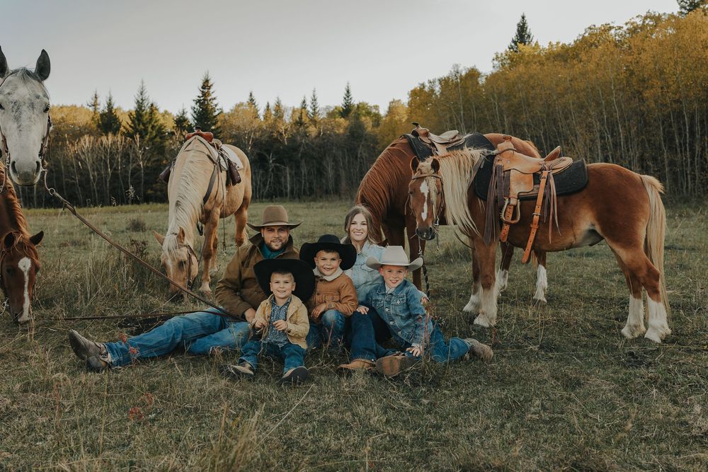 Family in cowboy hats sitting in a field with two horses and autumn trees in the background.