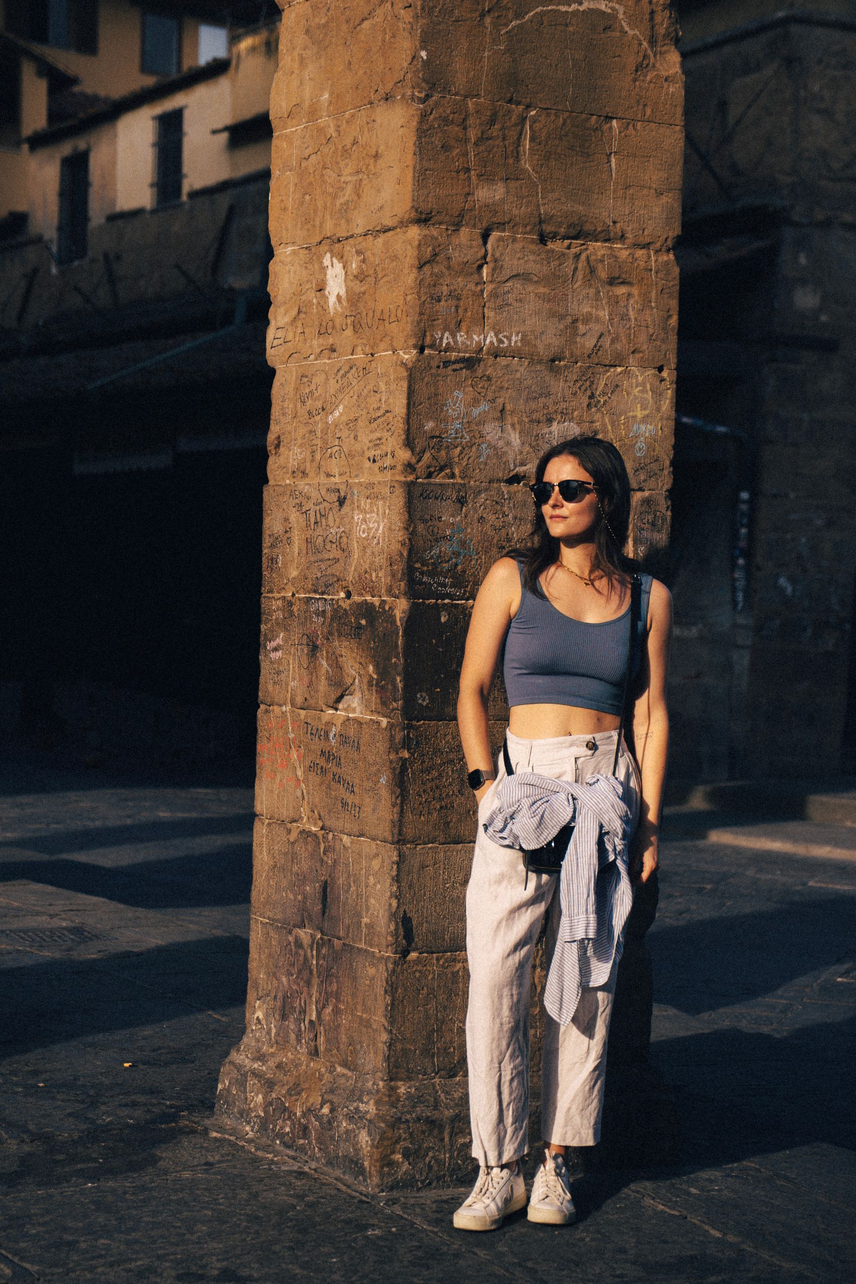 Trendy person in sunglasses and casual wear leaning against a rustic stone pillar in urban setting.