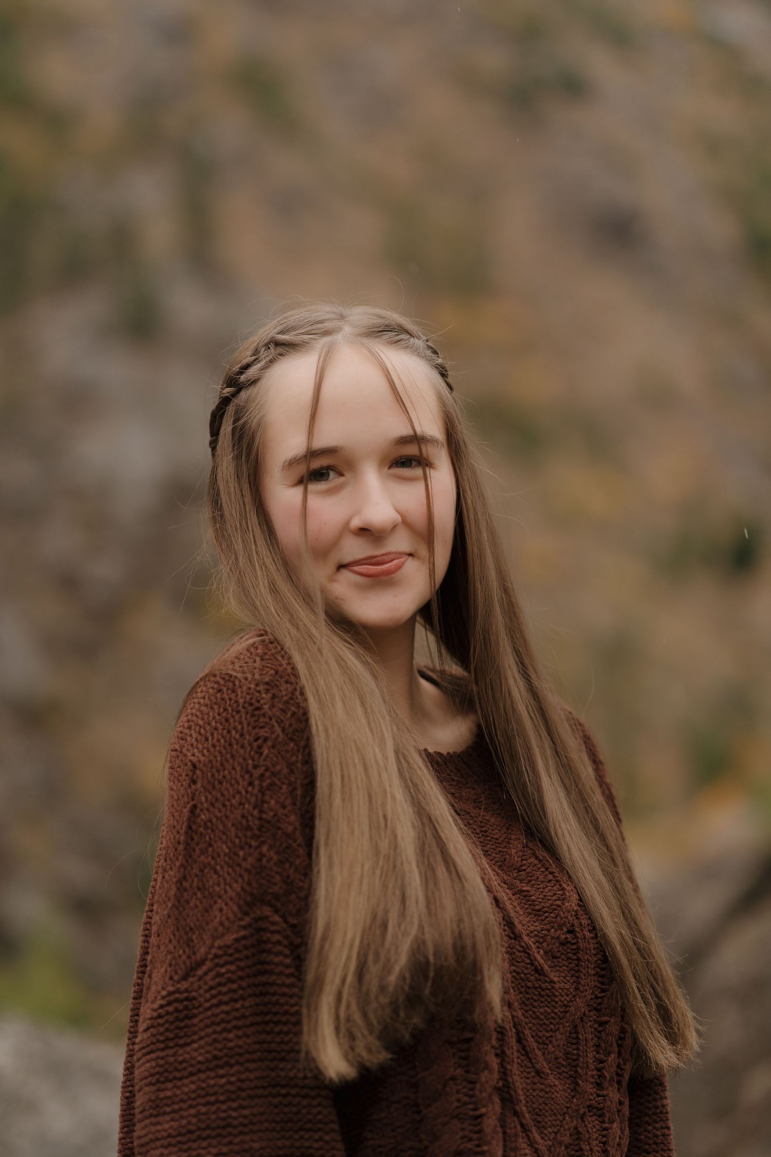A person with long blonde hair wearing a brown sweater smiles while posing against a blurred natural background.