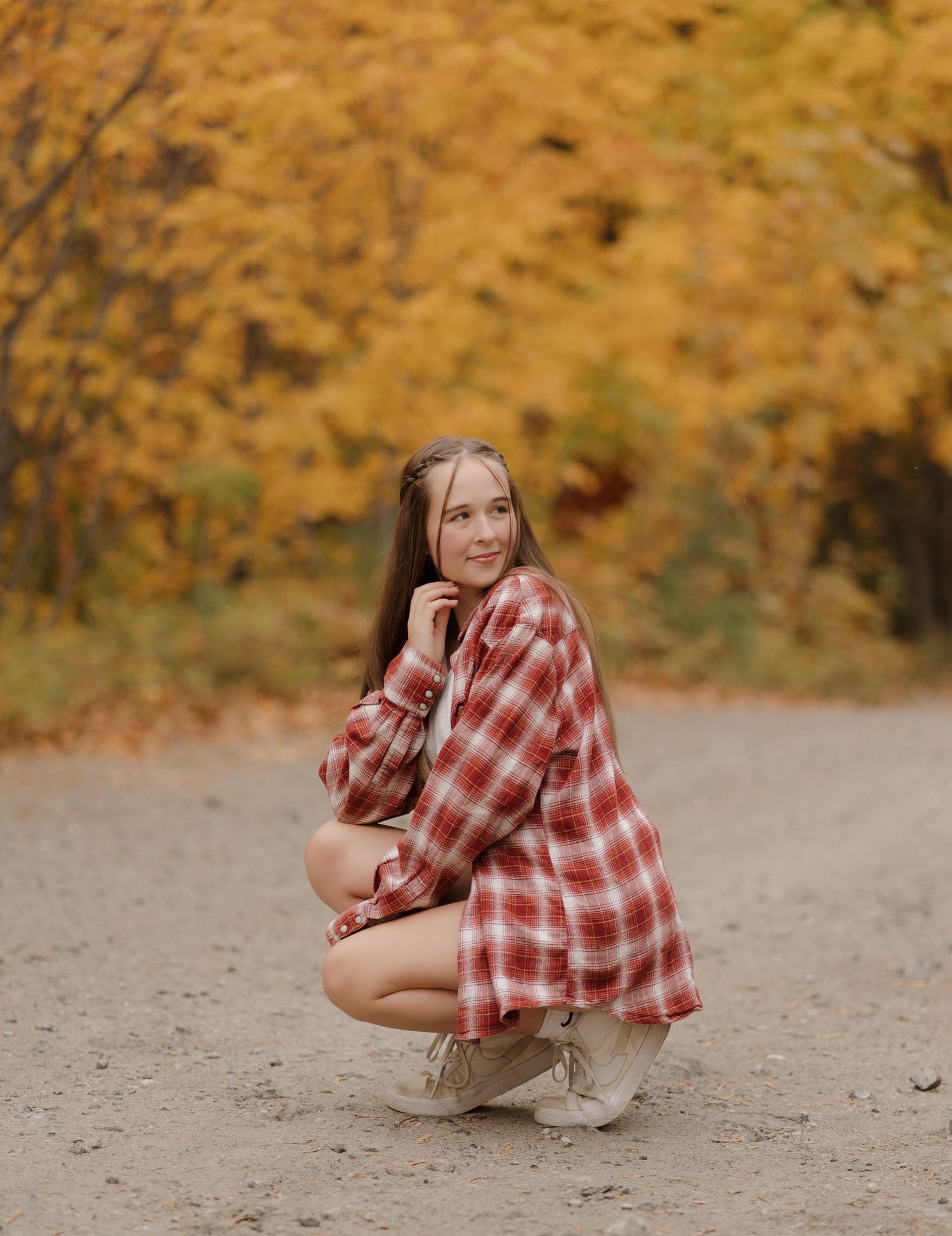 A person in a red plaid shirt and khaki pants crouches on a gravel path surrounded by autumn foliage.