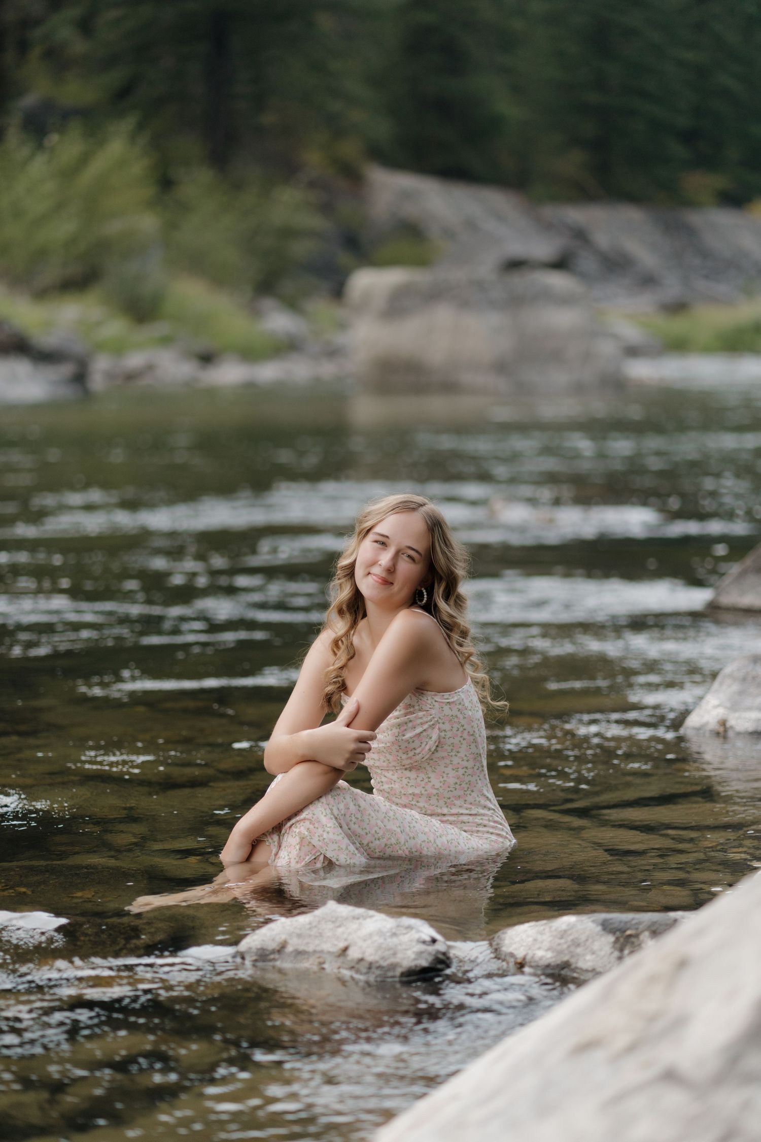 A person in a white dress reclines on rocks by a flowing river in a serene natural setting.
