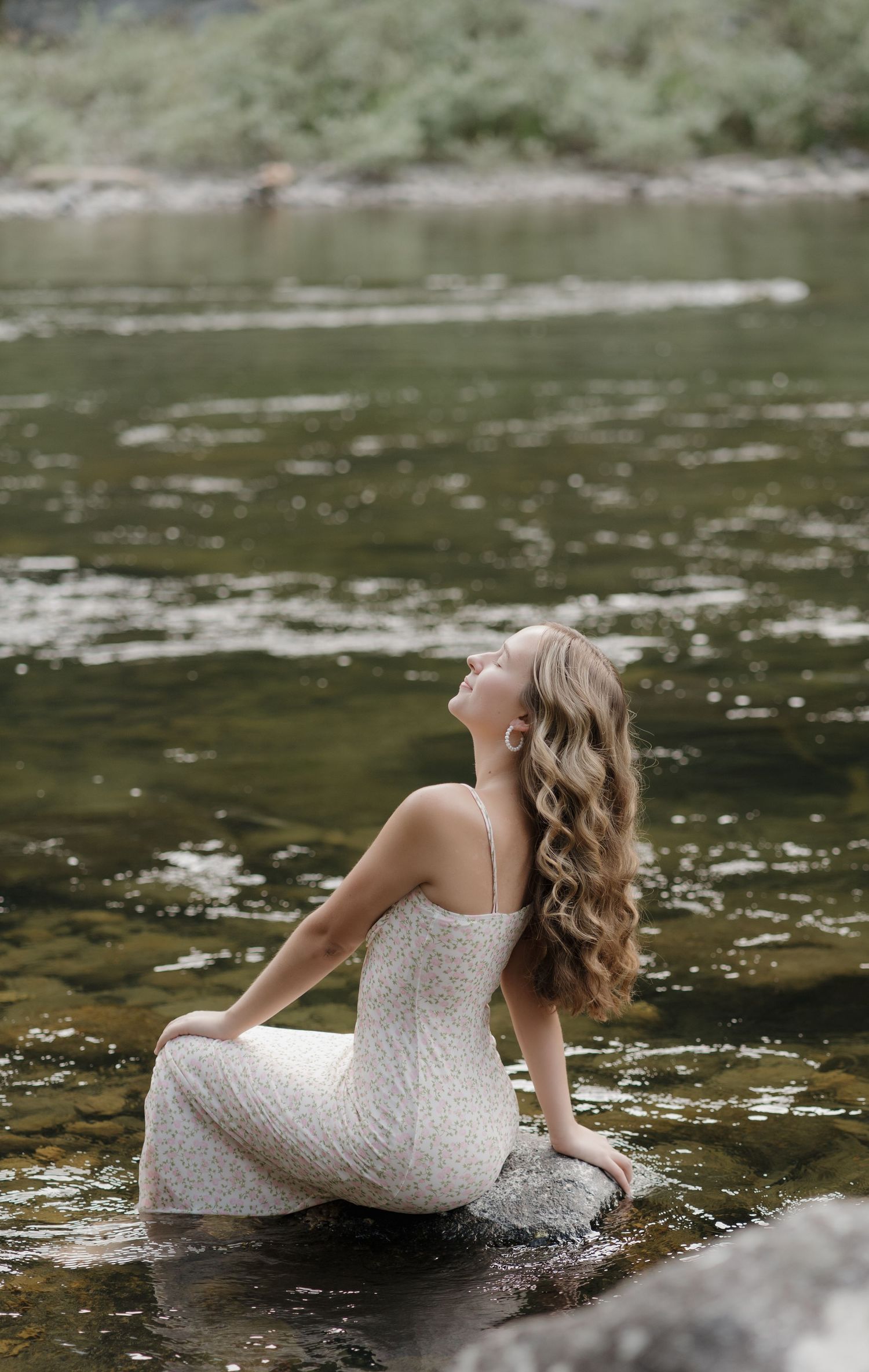 Person in white lace dress sits on rock in river with flowing water and greenery in background.