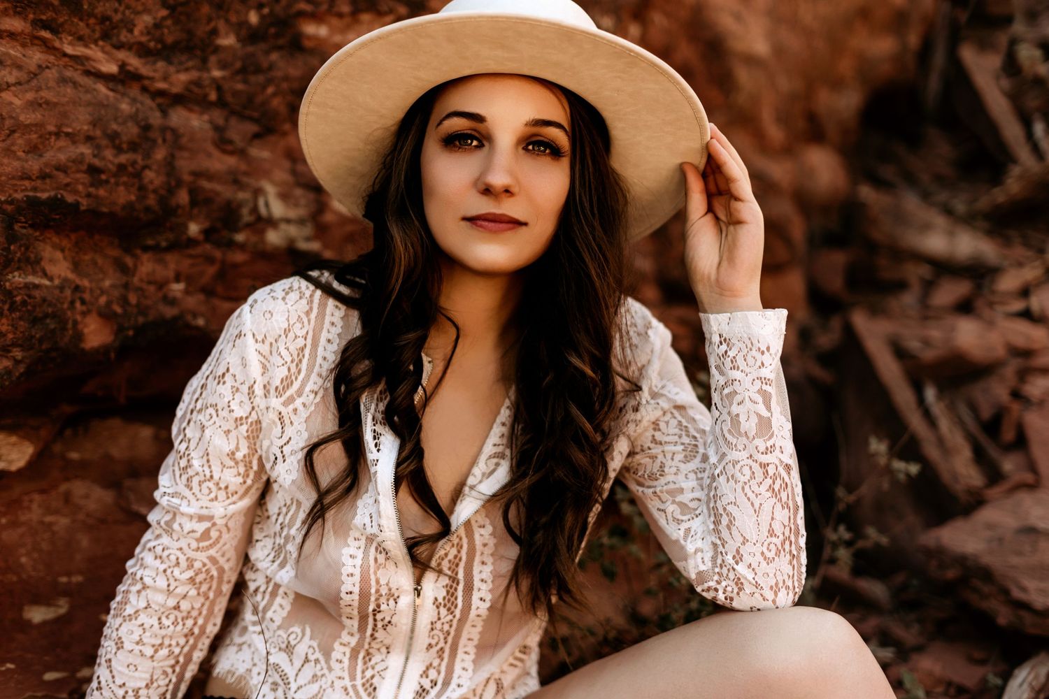 Portrait in desert setting with white lace top and wide brim hat against red rocks.