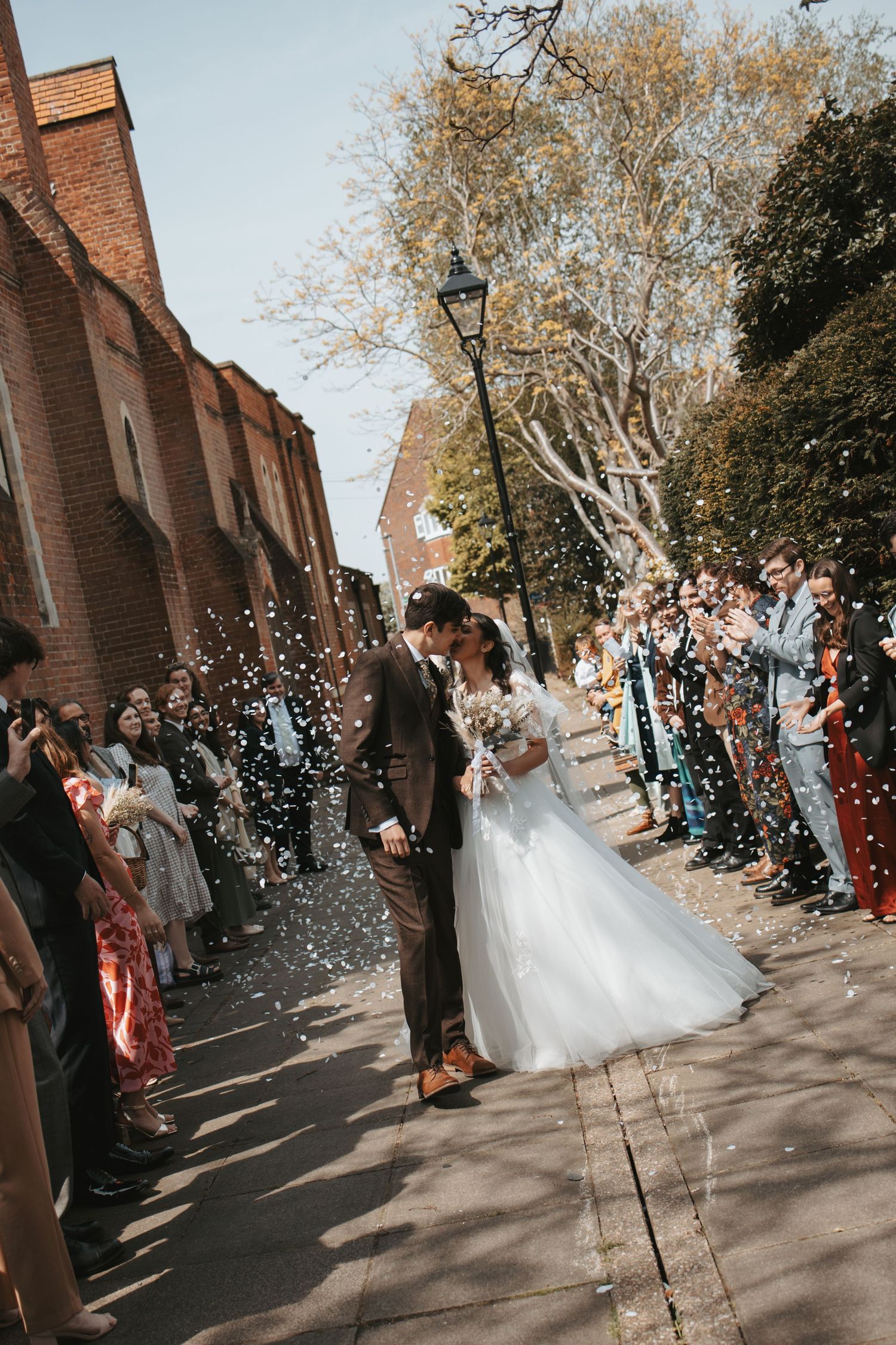 Wedding party exit with confetti shower outside a brick church on a sunny day.