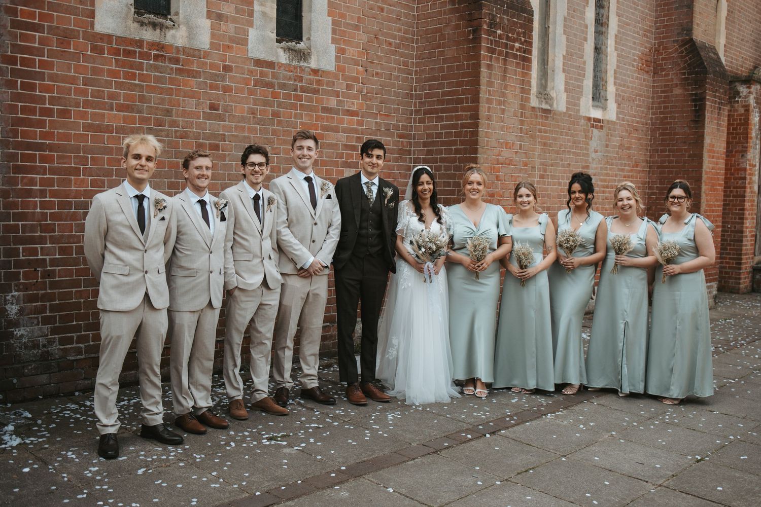 Wedding party in light gray attire poses along brick wall for formal group photo.